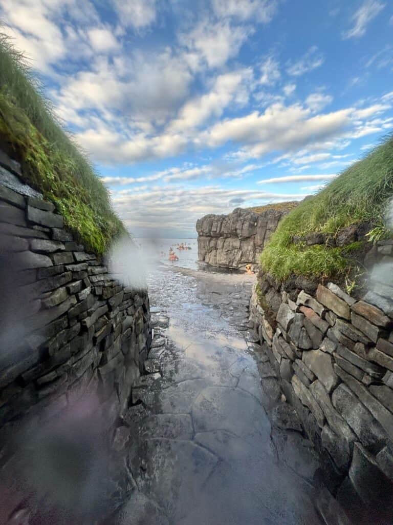 Green moss covered rock walls and water from Sky Lagoon with white fluffy clouds and teens bathing in Iceland