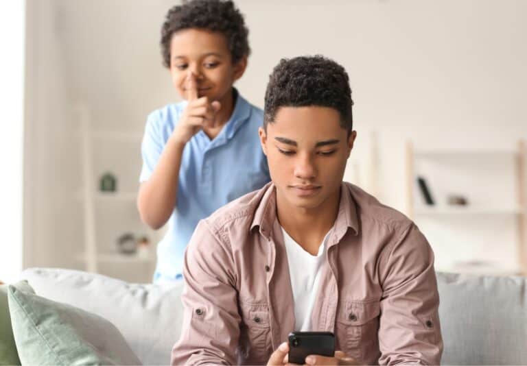 A young boy is saying shhh behind a teenage boy who he is about to prank