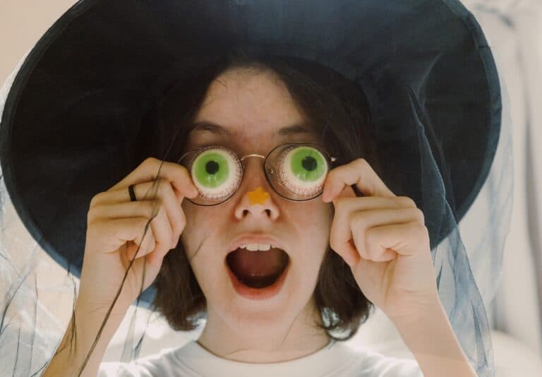 portrait of a teenage girl in a witch hat holding spooky googly eyes up to her eyes