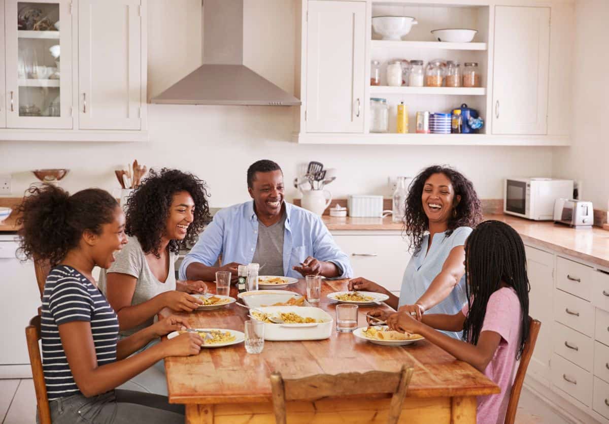 A smiling Black family with a mom, dad and three teenage girls sitting and laughing around the kitchen table