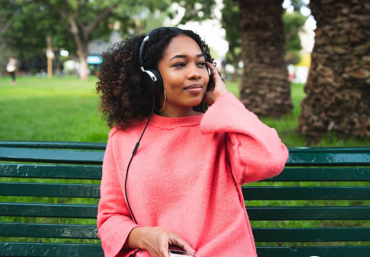 A Black teenager sitting on a park bench wearing headphones