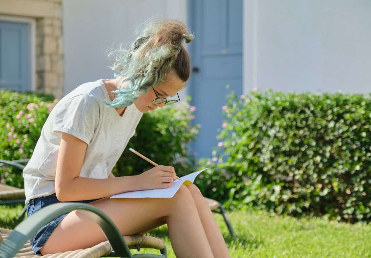 A teenager writing a gratitude letter on a wicker chair in a yard