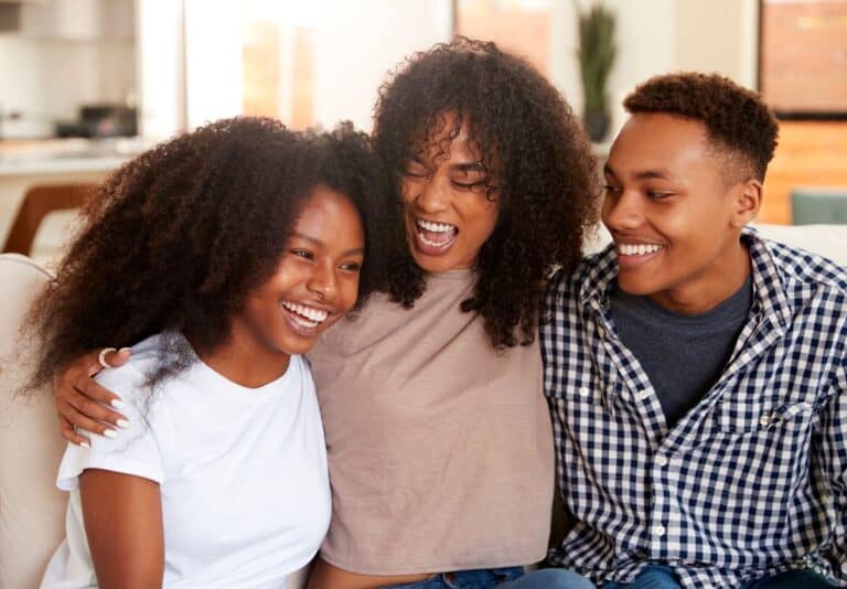 A Black mom with her teenage son and daughter sitting on the couch smiling, with their arms around each other