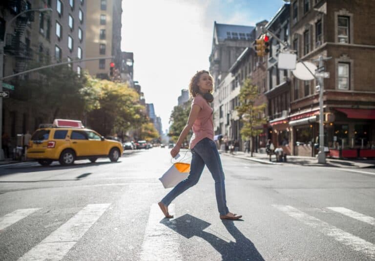 A teenage girl crossing the street in New York City holding a shopping bag.