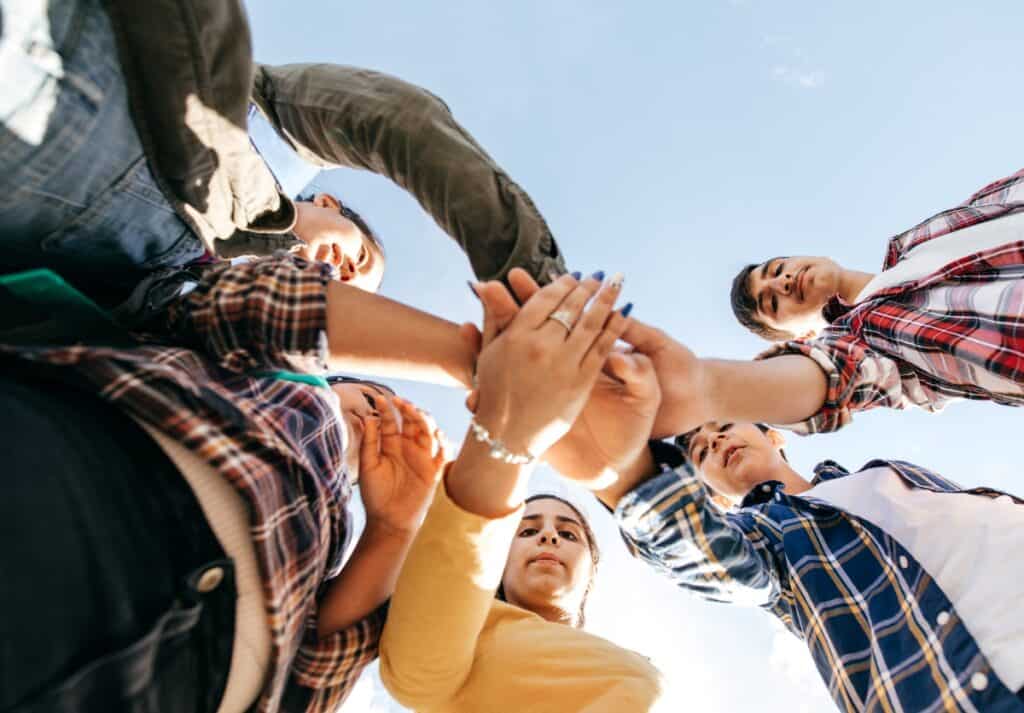 A group of teenagers putting their hands together in a circle. The shot is looking from the group up.