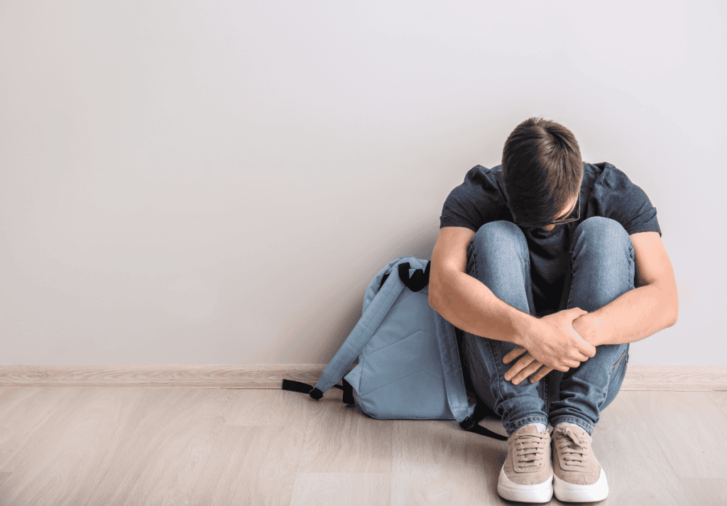 A bullied teen boy is sitting on the floor with his head on his knees.