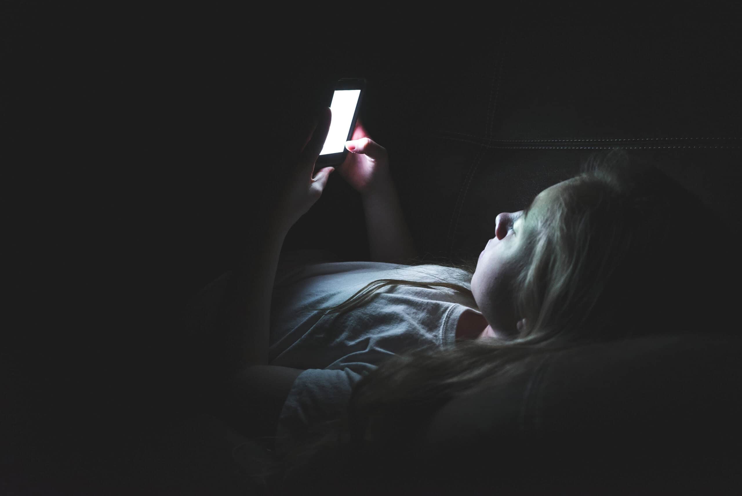 A teen in a dark room doomscrolling on their phone.